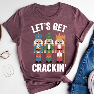 Puede incluir: Camiseta burdeos con la frase "LET'S GET CRACKIN'" sobre tres figuras de cascanueces de dibujos animados. Los cascanueces visten uniformes azules, verdes y rojos. La camiseta se combina con vaqueros azules y zapatillas blancas.