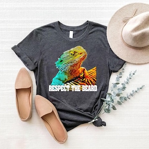 May include: Dark gray t-shirt featuring a colorful graphic of a bearded dragon with the text "Respect the Beard." The shirt is paired with beige flats and a beige hat.