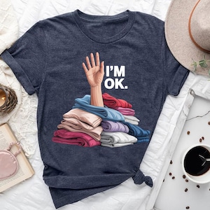 May include: Navy blue t-shirt with the text "I'M OK." above a graphic of a hand emerging from a pile of folded clothes in various colors. The shirt is knotted at the bottom.