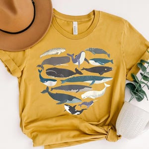 May include: Mustard yellow t-shirt featuring a heart-shaped arrangement of various whale illustrations in shades of gray, blue, and white. The shirt is made of soft material and is suitable for casual wear. A brown hat and white mug are also in the image.