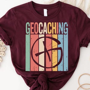 May include: A maroon t-shirt featuring the word "GEOCACHING" in a retro font above a circular graphic with a compass design. The graphic is set against a backdrop of vertical stripes in various colors, including teal, orange, and yellow.