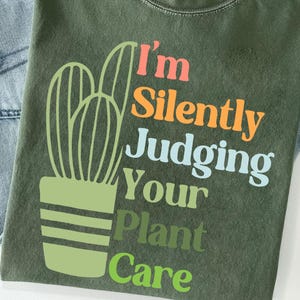 May include: Olive green t-shirt with a cactus graphic and the text "I'm Silently Judging Your Plant Care" in various colors. The shirt is folded on top of blue jeans.