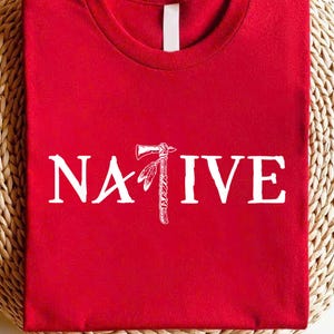 May include: A red t-shirt with the word "NATIVE" printed in white, featuring a Native American tomahawk graphic. The shirt is folded and displayed on a woven surface.