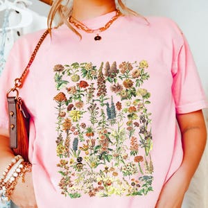A person wearing a pink t-shirt with a floral print, standing in front of a clothing rack with various garments hanging.
