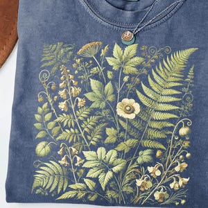 a blue t-shirt with a detailed embroidered design of various plants and flowers, including ferns, flowers, and leaves, creating a lush and vibrant floral pattern.