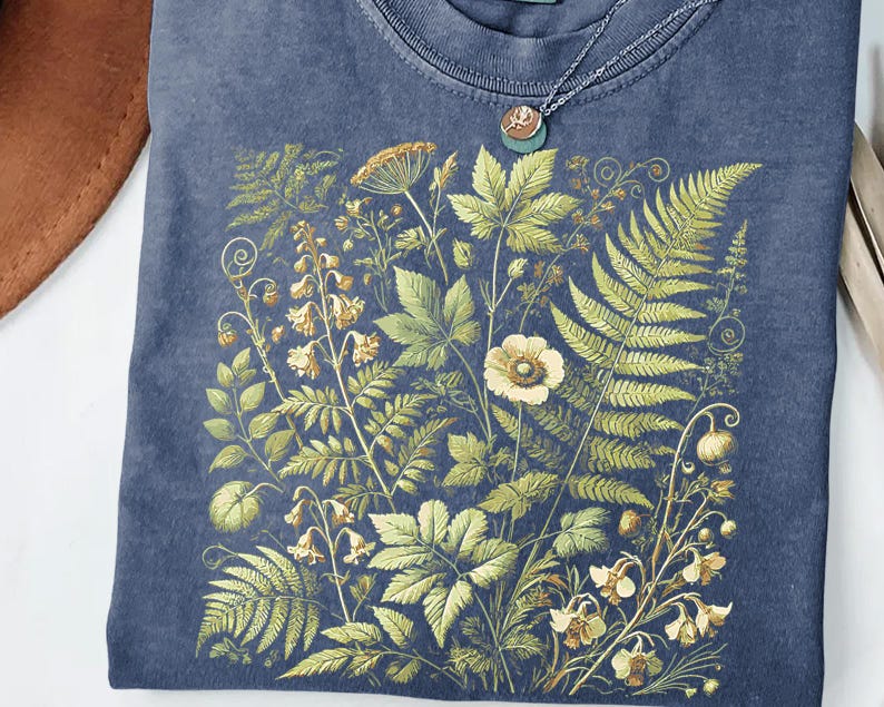 a blue t-shirt with a detailed embroidered design of various plants and flowers, including ferns, flowers, and leaves, creating a lush and vibrant floral pattern.