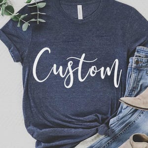 May include: Navy blue t-shirt with the word "Custom" in white script. The shirt is paired with blue jeans and tan ankle boots. A sprig of greenery is in the upper left corner.