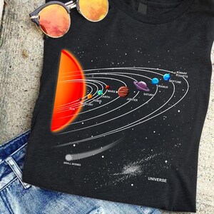 May include: A black t-shirt featuring a graphic of the solar system with a large red sun, orbiting planets labeled Mercury, Venus, Earth, Mars, Jupiter, Saturn, Uranus, Neptune, and Dwarf Planets, plus a comet labeled Small Bodies and the word Universe.