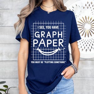 Puede incluir: Camiseta azul marino con un gráfico blanco de papel cuadriculado y el texto "I SEE, YOU HAVE GRAPH PAPER. YOU MUST BE 'PLOTTING SOMETHING'". El diseño incluye una cara sonriente.