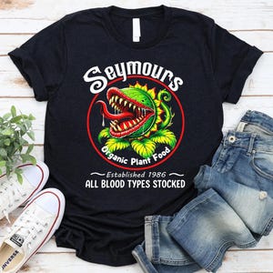 May include: Black t-shirt featuring a graphic of a carnivorous plant with the text "Seymours Organic Plant Food". The design includes a red circle and the words "Established 1986" and "All Blood Types Stocked".