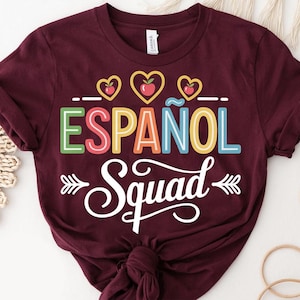 May include: Burgundy t-shirt with the words "ESPAÑOL Squad" in colorful letters and a white script font. The design includes heart-shaped graphics with apples. A casual, short-sleeved tee for Spanish language enthusiasts.