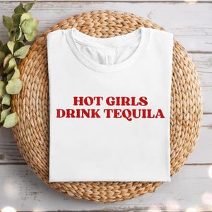 May include: A white t-shirt with the red text "HOT GIRLS DRINK TEQUILA" is displayed on a woven, circular placemat. The shirt is folded neatly, showcasing the bold statement. Green foliage accents the scene.