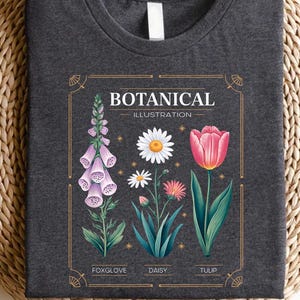 May include: Dark gray t-shirt featuring a botanical illustration. The design includes a foxglove, daisy, and tulip, with the words "Botanical Illustration" above the flowers. The shirt has a classic crew neck.