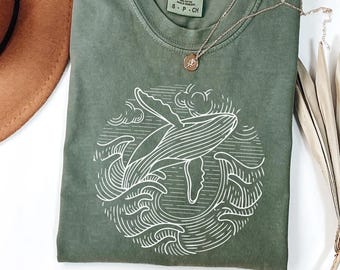 May include: Olive green t-shirt featuring a white line drawing of a whale breaching the ocean waves. The design is within a circular frame. A gold necklace with a small pendant rests on the shirt.