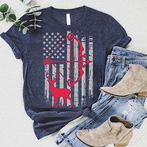 May include: Navy blue t-shirt featuring a distressed American flag design with a red bow and arrow, and a red deer silhouette. The shirt is paired with blue jeans and tan ankle boots.