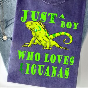 May include: A purple t-shirt featuring a bright green iguana illustration in the center. The text printed in green reads JUST A BOY WHO LOVES IGUANAS, with small lizard silhouettes integrated into the lettering. The shirt has a crew neckline.