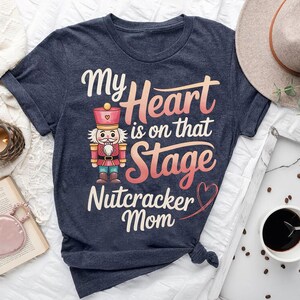 May include: Navy blue t-shirt with the text "My Heart is on that Stage Nutcracker Mom" and a cartoon nutcracker illustration. The design features a pink and orange gradient, a heart, and a decorative font.