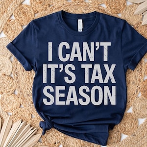 May include: Navy blue t-shirt with the text "I CAN'T IT'S TAX SEASON" in distressed white lettering. The shirt is displayed on a woven, beige, circular rug with a natural fiber texture. A few dried leaves are placed on the right side of the shirt.