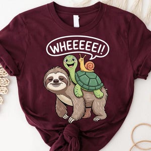 May include: Burgundy t-shirt featuring a cartoon sloth with a turtle and snail. The turtle is riding on the sloth's back, and the snail is on the turtle. The text "WHEEEEE!!" is in a white speech bubble.