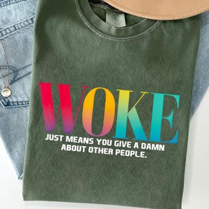 May include: An olive green t-shirt with the word "WOKE" in a rainbow gradient across the front. Below the word is the text "JUST MEANS YOU GIVE A DAMN ABOUT OTHER PEOPLE." The shirt is laid on top of blue jeans and a tan hat.
