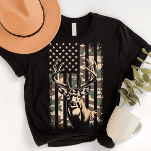 a black t-shirt with a deer graphic and an American flag design, along with a cowboy hat, a mug, and some greenery.