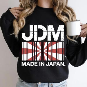 Peut inclure: Sweat-shirt noir avec le texte blanc "JDM" au-dessus d'un graphique du drapeau japonais et les mots "MADE IN JAPAN." Le sweat-shirt est de style col rond.