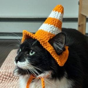 Könnte beinhalten: Eine schwarz-weiße Katze trägt einen handgefertigten gehäkelten Verkehrskegelhut mit orangefarbenen und weißen Streifen. Der Hut hat eine spitze Spitze und ist mit orangefarbenen Bändern befestigt. Die Katze liegt auf einer rosa strukturierten Oberfläche.