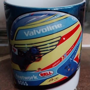 Puede incluir: Una taza de cerámica blanca con un diseño de casco de carreras azul, amarillo y rojo. El casco tiene las palabras "Valvoline", "Bell Helmets", "Speed", "Finetwork Boss" y "Arcand" en él.
