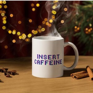 Op de afbeelding: Witte keramische koffiemok met de tekst "INSERT CAFFEINE" in een gepixelde, geglitchte lettertype in paars en blauw.