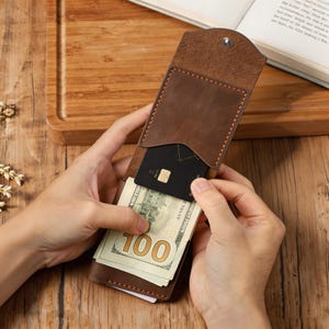 May include: A brown leather wallet with a snap closure, featuring a vertical pocket containing a black credit card and a stack of one hundred dollar bills. The wallet has visible stitching along the edges and a textured flap.