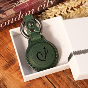 May include: A dark green leather keychain holder with a circular design and a metal ring attachment. The front surface features a black engraved symbol consisting of a heart shape combined with a cross. The item is placed inside a white gift box.