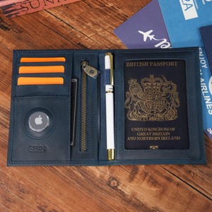 May include: Open navy blue leather travel wallet with orange card slots, a zippered pocket, and a pen. A British passport is visible. The wallet has an RFID protection logo and an Apple AirTag.
