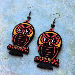 May include: A pair of black wooden earrings with a red and yellow owl design. The owls have large eyes and are perched on a branch.