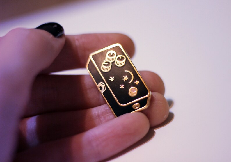 May include: Gold and black enamel pin shaped like a guitar pedal with a crescent moon, stars, and three knobs.