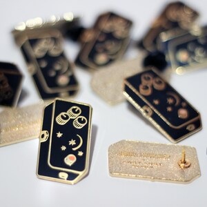 May include: A close-up of a black and gold enamel pin featuring a stylised illustration of a musical pedal with three knobs, stars, and a crescent moon. The pin is on a white surface with other pins and a backing card that reads "Lemon Squeezy" with a website address.