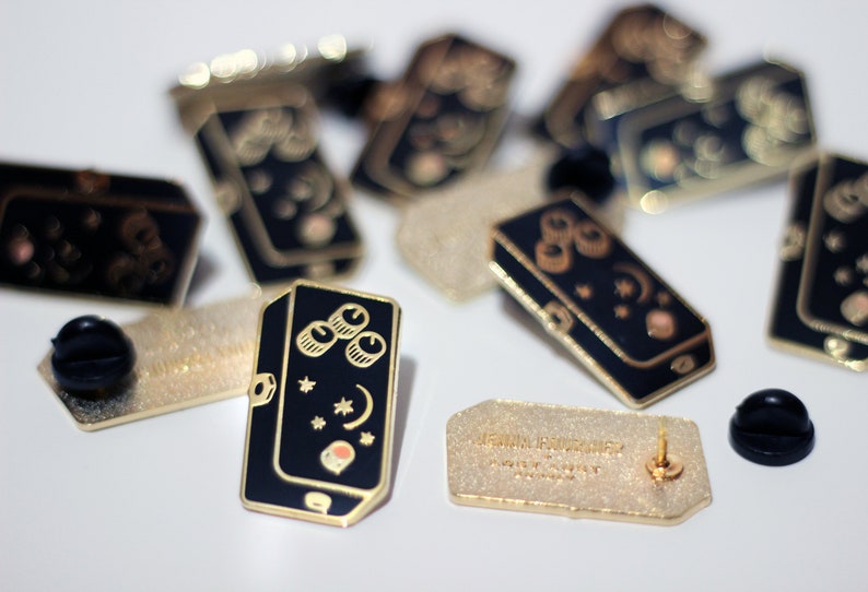 May include: A close-up of a black and gold enamel pin featuring a stylised illustration of a musical pedal with three knobs, stars, and a crescent moon. The pin is on a white surface with other pins and a backing card that reads "Lemon Squeezy" with a website address.