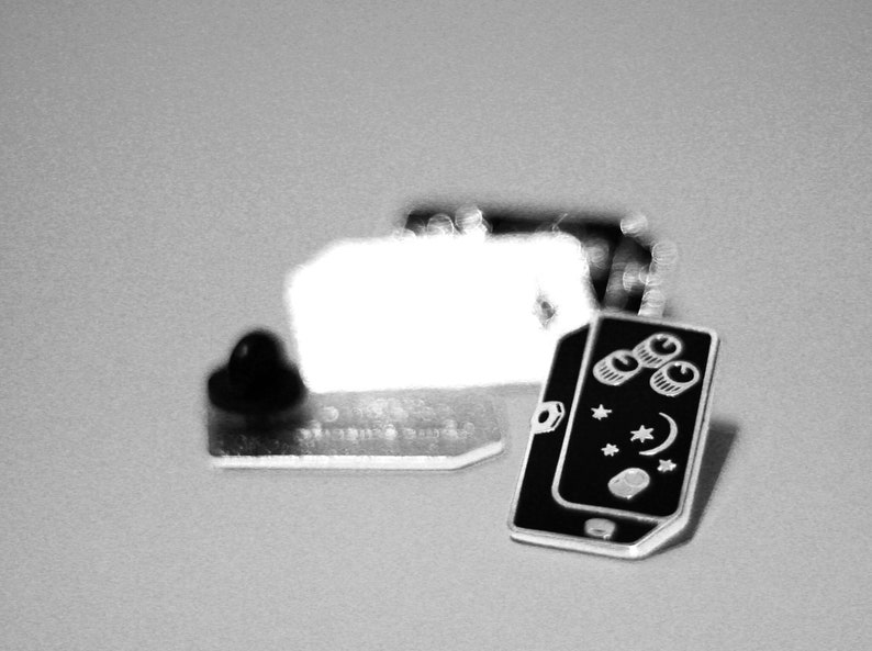 May include: Black and white enamel pin of a mobile phone with three knobs and a crescent moon and stars design.