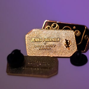 May include: Gold and black enamel pin featuring the text "Jenna Fournier Lost Lusty Supply" and a graphic of a camera with a star and moon.