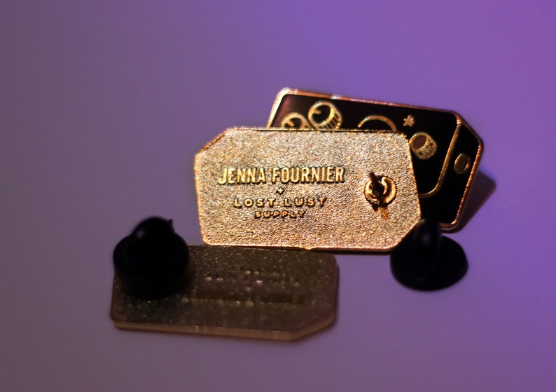 May include: Gold and black enamel pin featuring the text "Jenna Fournier Lost Lusty Supply" and a graphic of a camera with a star and moon.