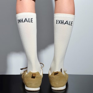 May include: A pair of white socks with black text. The left sock says "INHALE" and the right sock says "EXHALE". The socks are worn with brown suede sneakers.