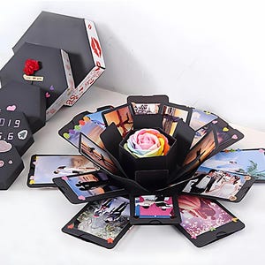 Puede incluir: Caja explosiva negra con fotos y una rosa arcoíris en el centro. La caja está abierta, revelando múltiples paneles fotográficos. Se ven cajas negras cerradas adicionales, una con una rosa roja y otra con la fecha "2019.6.6".