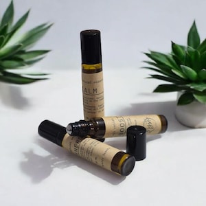 May include: An assortment of essential oil roller bottles with black caps and tan labels. The labels read "Calm", "Boost", and "Energy". Two green succulent plants in white pots are in the background.