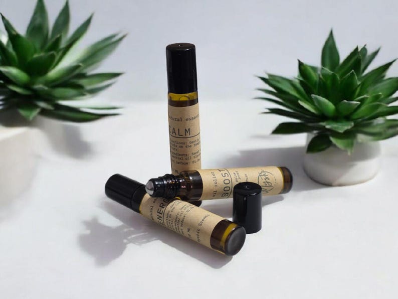 May include: An assortment of essential oil roller bottles with black caps and tan labels. The labels read "Calm", "Boost", and "Energy". Two green succulent plants in white pots are in the background.