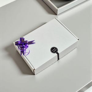 May include: A white rectangular gift box with a purple ribbon and bow. A black sticker with the words "hand made" is on the box. The box is on a gray surface.