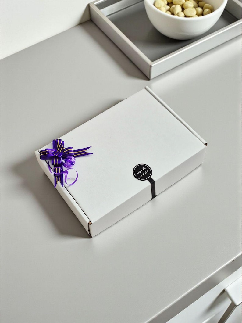May include: A white rectangular gift box with a purple ribbon and bow. A black sticker with the words "hand made" is on the box. The box is on a gray surface.
