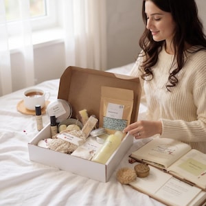 May include: A white gift box filled with self-care products, including essential oils, bath salts, and a loofah. The box is open, revealing various items in a natural color palette. A woman is seen opening the box on a bed with a book and a cup of tea nearby.
