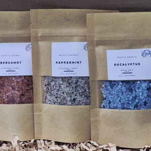 May include: Three kraft paper pouches with clear windows, each containing mineral soap. The pouches are labeled "Bergamot," "Peppermint," and "Eucalyptus." The soap crystals are red, gray, and blue, respectively. The text "Mystic Scents" is visible.