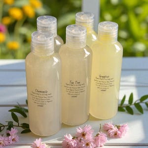 May include: Five clear plastic bottles of body wash with flip-top caps. Each bottle has a label with a different scent: Chamomile, Tea Tree, and Grapefruit. The bottles are arranged on a white surface with pink flowers.