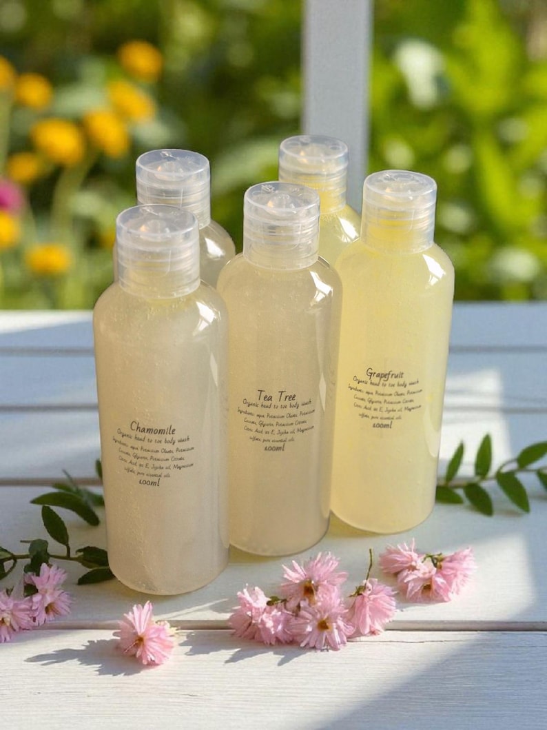 May include: Five clear plastic bottles of body wash with flip-top caps. Each bottle has a label with a different scent: Chamomile, Tea Tree, and Grapefruit. The bottles are arranged on a white surface with pink flowers.