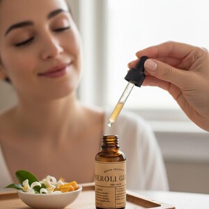 May include: A brown glass bottle of "NEROLI GLOW" serum with a dropper, held above the bottle. A small bowl with orange slices and white flowers sits on a wooden tray. The serum is being applied to a person's face.